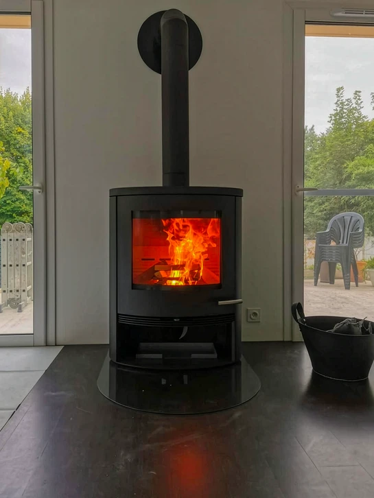 Intérieur chaleureux avec poêle à bois en fonctionnement, flammes visibles à travers la vitre, réserve de bûches intégrée dans la partie basse